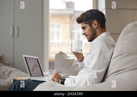Young focused european businessman drinking tea or coffee from cup and watching business graph on laptop. Concept of modern successful man. Bearded st Stock Photo