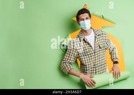 Smiling young casual white man looking out from a torn paper hole background wearing protective face mask celebrating birthday party Stock Photo