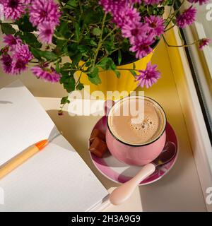 Coffee cup chocolate notebook beautiful pink autumn Chrysanthemum flowers in yellow pot on window. Cozy home Stock Photo