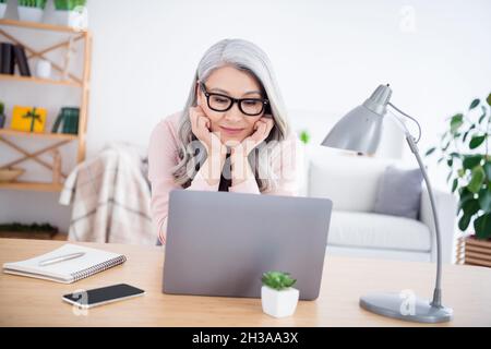 Photo of charming positive aged person sit behind desk hands on cheeks look interested laptop video indoors Stock Photo