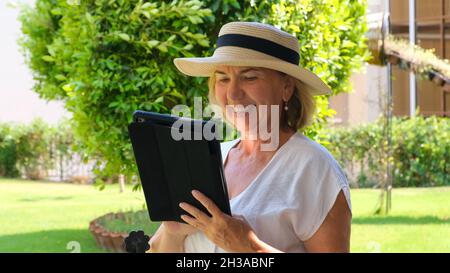 Senior female blonde businesswoman in hat uses tablet pc, digital tablet for business work or study in her own green garden. woman aged 50-55. The Stock Photo