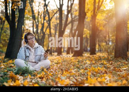Caucasian middle aged woman in earphones listening to music, meditation app on smartphone and meditating in lotus pose at autumn park. Meditation app Stock Photo