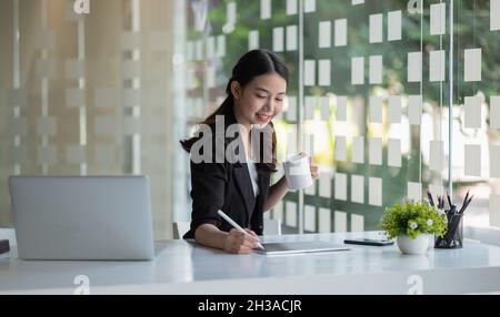 Happy beautiful Young Asian business woman working with digital tablet while sitting at table in co-working office. Freelance Work, smiling, positive Stock Photo