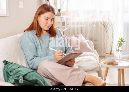 Charming cheerful woman wears pajamas, sit on sofa hold tablet takes notes in diary data work at home indoors Stock Photo