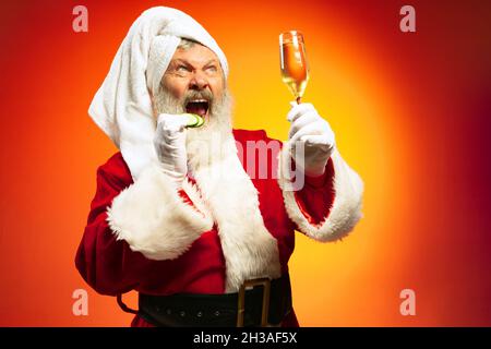 Funny senior man wearing white towl and costume of Santa Claus congrats everybody with New Year 2022 isolated on red yellow background in neon. Stock Photo