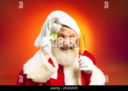 Funny senior man wearing white towl and costume of Santa Claus congrats everybody with New Year 2022 isolated on red yellow background in neon. Stock Photo