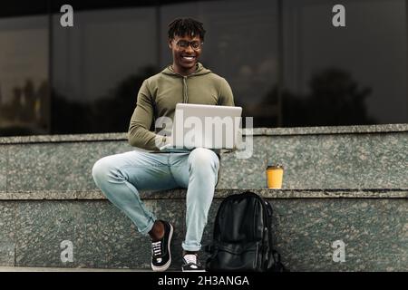 Smiling african man, using laptop sitting on city stairs, making notes, watching webinar, engaging in online lesson, motivated positive student studyi Stock Photo