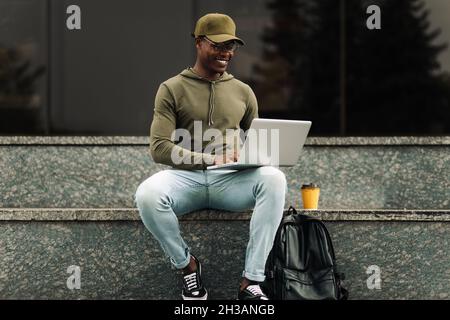 Smiling african man, using laptop sitting on city stairs, making notes, watching webinar, engaging in online lesson, motivated positive student studyi Stock Photo