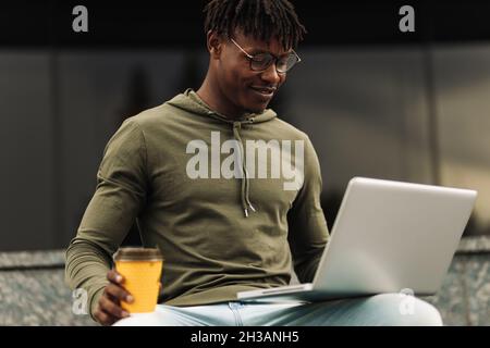 Young African American male businessman, wearing glasses working online with laptop sitting on a bench holding a takeaway coffee cup near his office b Stock Photo