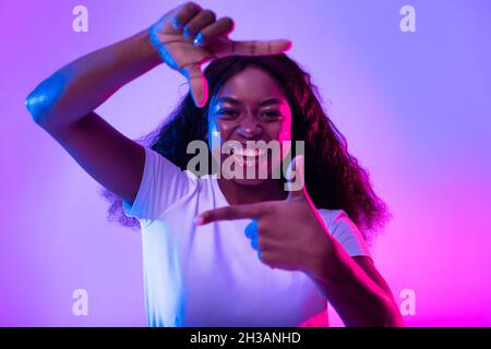Happy black woman making picture frame with fingers, looking at camera and smiling for photo in neon light Stock Photo