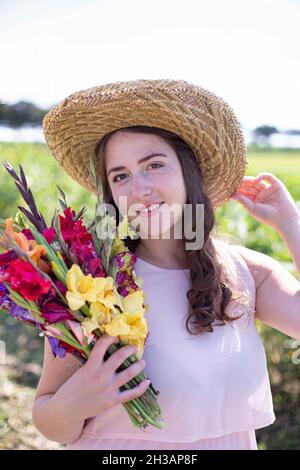 White young woman with long hair in light dress in straw hat with bright bouquet of flowers in the countryside Stock Photo
