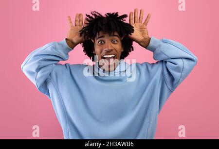 Cool African American teen guy making silly gesture with his hands, grimacing and having fun on pink studio background Stock Photo