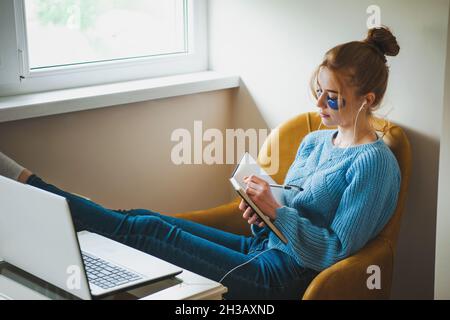Woman taking notes while participates at online classes sitting relaxed on armchair. Freelance work. Stock Photo