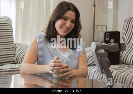 Young girl blogger smiles contentedly at camera while talking to her followers through a live video for her social media while enjoying her favorite m Stock Photo