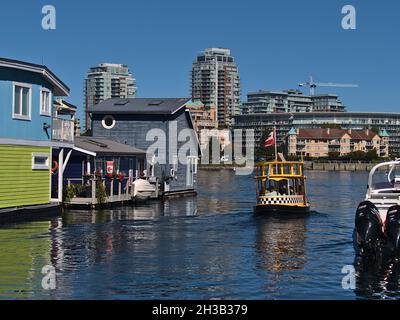 Small Victoria Harbour Ferry departing from Fisherman's Wharf with colorful floating houses and skyline in background on sunny day. Stock Photo