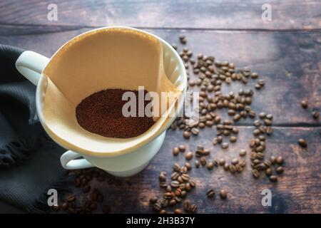 Ground coffee in a paper filter bag and in a porcelain holder on a mug for an aromatic drip brewed hot drink, rustic wooden table with some beans, cop