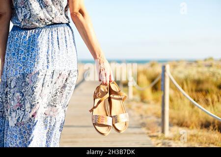 Middle aged woman walking on the beach with sandals in her hand. She is wearing a flowery summer dress.