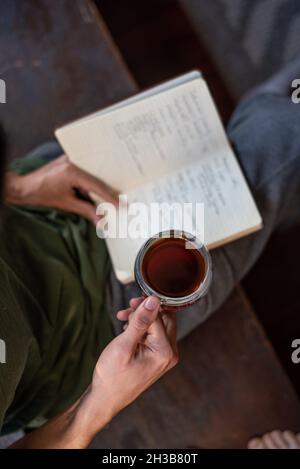 Aerial view of mixed race man with black leather-bound journal and clear mug of black coffee, lounging in gray sweats with green top. Stock Photo