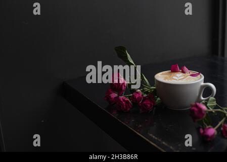 Rose cardamom latte with latte art in white latte mug on dark coffee shop counter garnished with rose petals and styled with pink roses. Stock Photo
