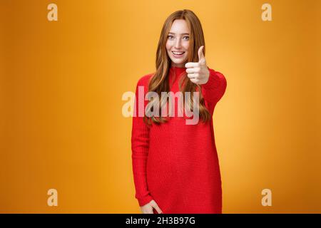 Nice job good. Portrait of delighted and energized confident ginger woman in red sweater pulling thumb up at camera and smiling, liking idea