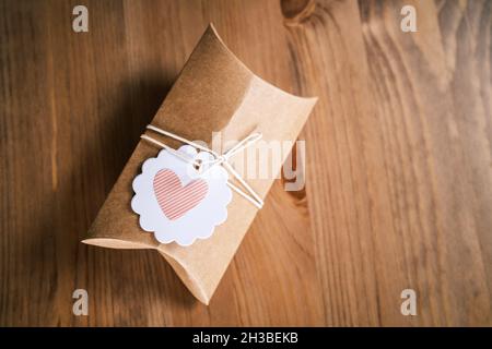 Small gift wrapped in craft paper on wooden background. Homemade present, heart shape on gift label. Sustainable gift wrapping using recycled paper. Stock Photo