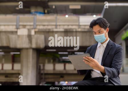 Asian businessman wearing face mask and sitting outdoors in city using digital tablet computer Stock Photo