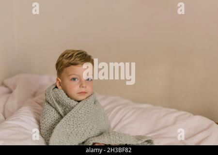 Toddler boy wrapped in grey blanket sitting on bed being ill Stock Photo