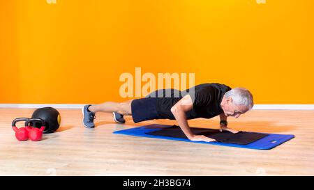 focused senior fit man doing push up workout exercise laying at the gym isolated on orange background. mature people fitness training indoor. elderly Stock Photo