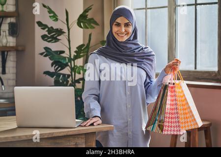 Elegant Muslim lady holding a bunch of gift bags and standing near a laptop Stock Photo
