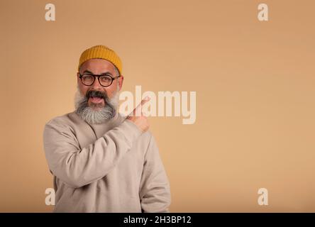 Emotional photo of a dejected bearded man opens his mouth wide, looks at the camera and points in the free space, says: Wow, that's something incredib Stock Photo