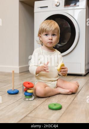 A small beautiful child is sitting on the floor of the house and playing with a wooden toy pyramid of colorful rings. Surprise Stock Photo