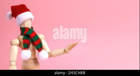 Wooden mannequin for art supplies store. Christmas banner with a moving doll made of wood in a striped scarf and Santa hat on a pink background. Stock Photo