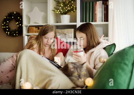 Two young sisters snuggling up on the sofa in a cozy living room at Christmas. Cute children using a tablet at home during winter break. Kids reading Stock Photo