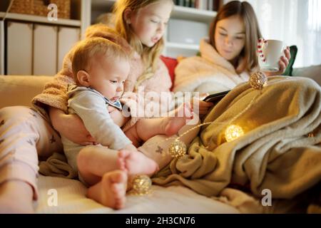 Two young sisters and their baby brother snuggling up on the sofa in a cozy living room at Christmas. Cute children using a tablet at home during wint Stock Photo
