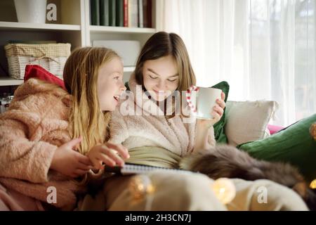 Two young sisters snuggling up on the sofa in a cozy living room at Christmas. Cute children using a tablet at home during winter break. Kids reading Stock Photo