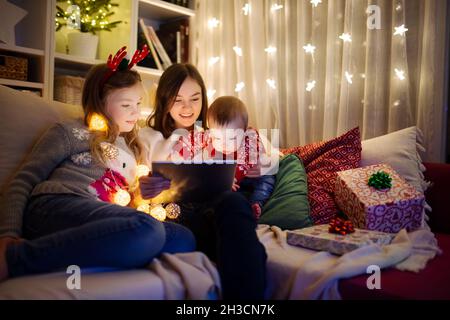 Two girls and a baby boy using a tablet pc at home in warm and cozy living room at Christmas. Family having online video call on Xmas eve. Winter even Stock Photo
