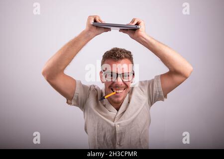 Young guy goes crazy in the workplace, stressed worker lifts the laptop and has pen in his mouth, angry millennial nerd maddened by stress Stock Photo