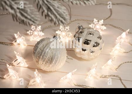 White with silver Christmas balls lie on a white wooden table along with a luminous garland. View through a pine branch. Closeup Stock Photo