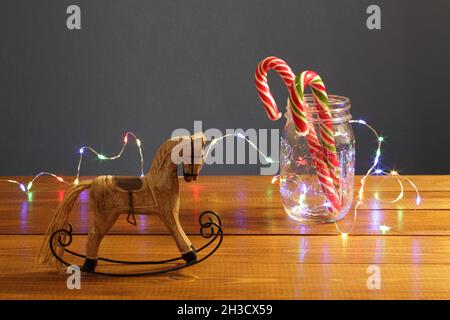 Christmas decoration - small wooden toy rocking-horse and caramel New Year's candies in a glass jar entwined with a luminous festive garland on a tabl Stock Photo