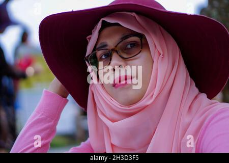 Close up portrait of beautiful young Asian muslim woman wearing hat, pink dress and hijab. Looking at camera with poker face expression. Stock Photo