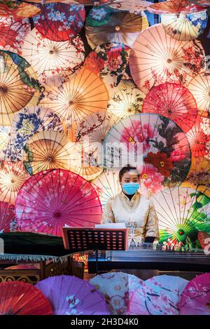 Chengdu, Sichuan province, China - Oct 24, 2021 : Young Chinese woman with a face mask playing Guzheng with chinese traditional umbrellas decoration in KuanZhai alley Stock Photo