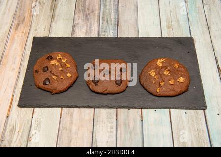 Chocolate chip cookies of different flavors with chocolate chips, pieces of nuts and pieces of dried fruits on black slate plate Stock Photo