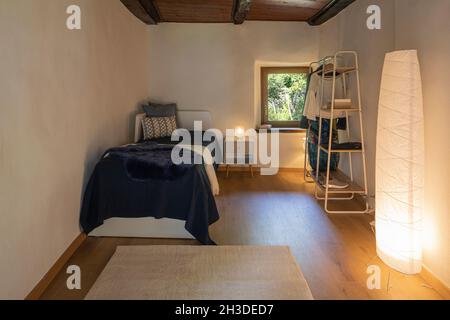 Front view bedroom with single bed, bedside table with a lamp above and a wardrobe on the right. There is a small window with a view of nature. Nobody Stock Photo
