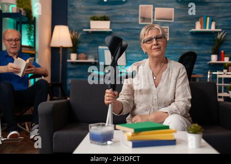Old woman holding crutches and looking at camera in interior space. Senior adult with handicap sitting on couch while retired man in wheelchair reading in living room background Stock Photo