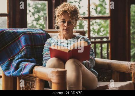 Pretty middle age woman relax at home sitting on an armchair reading a book enjoying indoor leisure activity. Adult female people comfortably sit down Stock Photo