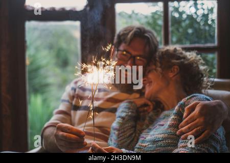 Couple celebrate alone at home with smile and fire sparkler sitting on the couch qieh outdoor nature view. Happy caucasian people in love and friendsh Stock Photo