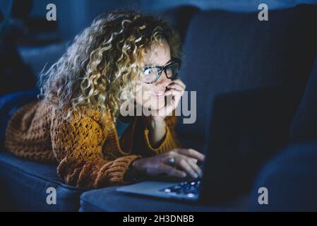 Attractive adult woman smile and use laptop computer and internet connection by night laying on the sofa at home. People addicted to technology and so Stock Photo