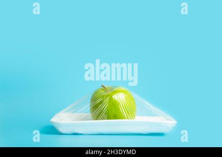 Fruits in plastic packaging from the supermarket are minimal. Apple in cellophane and non-degradable plastic on a blue background. Biodegradable product packaging, environmental protection, nature pollution. High quality photo Stock Photo