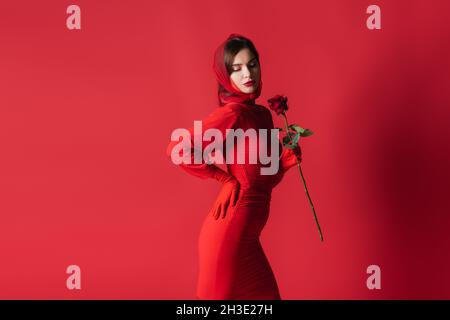 elegant young woman in dress holding rose while posing with hand on hip on red Stock Photo