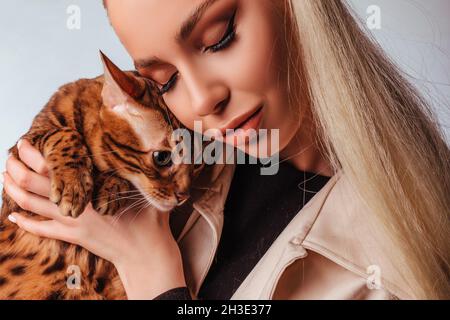 Portrait of an attractive cute young blonde woman with a bengal cat. Beautiful girl, looks at the cat, elegant hairstyle, impudent makeup, White Stock Photo